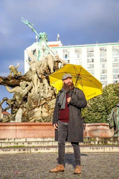 Bearded man in a hat holding a yellow Walkative umbrella, standing in front of the Neptune Fountain in Berlin.