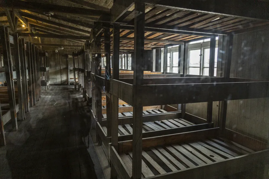 Interior of a wooden barrack with multi-tiered bunk beds, long rows extending into the dimly lit space. Windows are visibl...