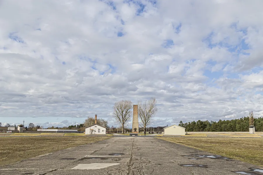 Concrete path leading to a tall, triangular brick monument flanked by two bare trees, with low buildings and fields under ...