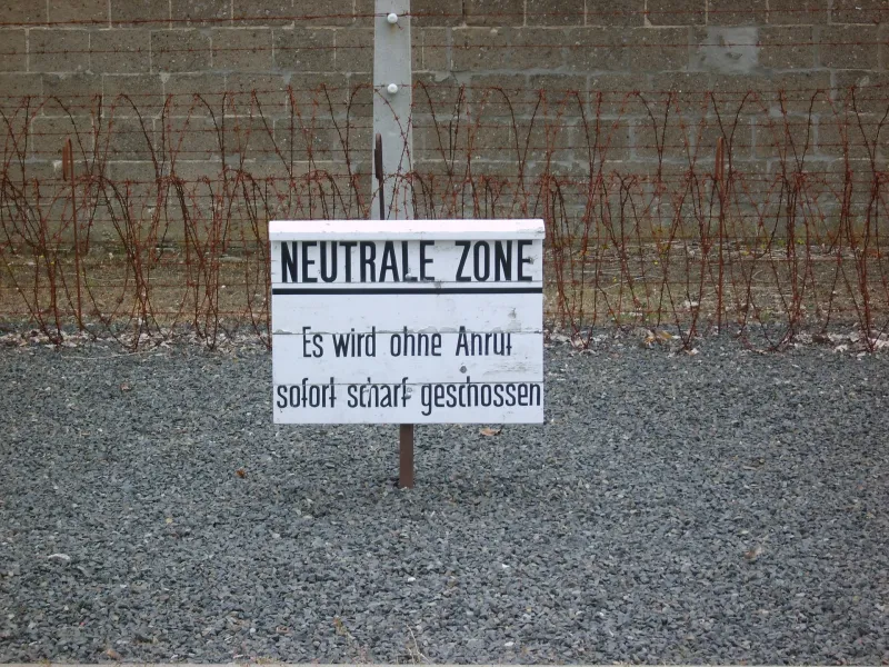 White sign with "NEUTRALE ZONE" and warning text in German, in front of barbed wire fence and concrete wall on gravel ground.