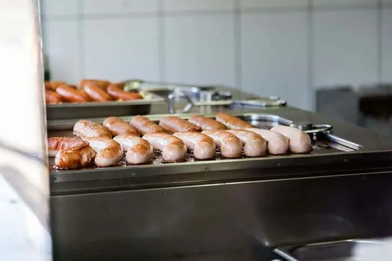 Sausages cooking on a metal griddle in a kitchen setting with white tiled walls. Metal tongs are visible.