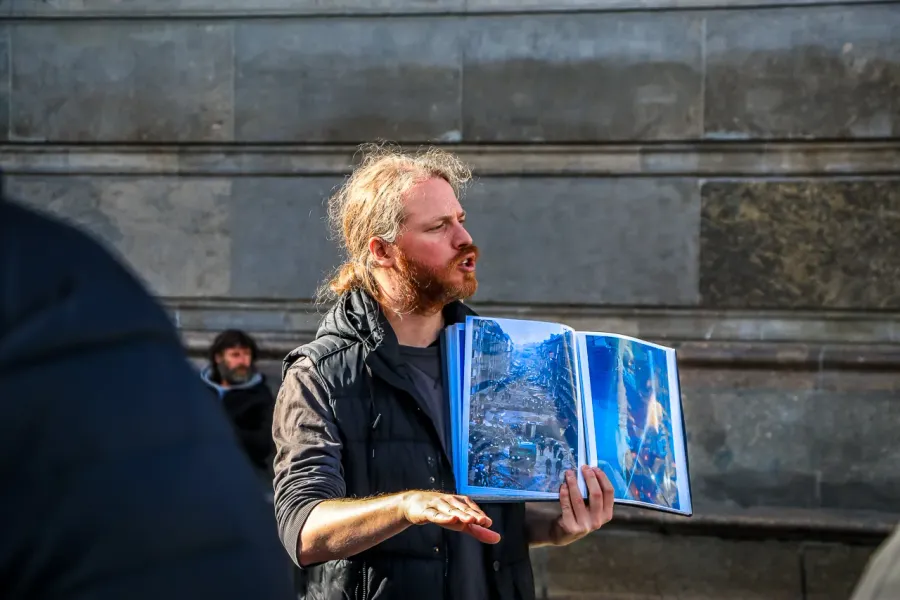 A tour guide in Berlin holds an open book showing a historical photo of a destroyed street while speaking to a group.