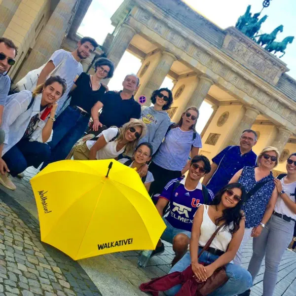 A diverse group of people posing with a yellow Walkative umbrella in front of the Brandenburg Gate in Berlin.