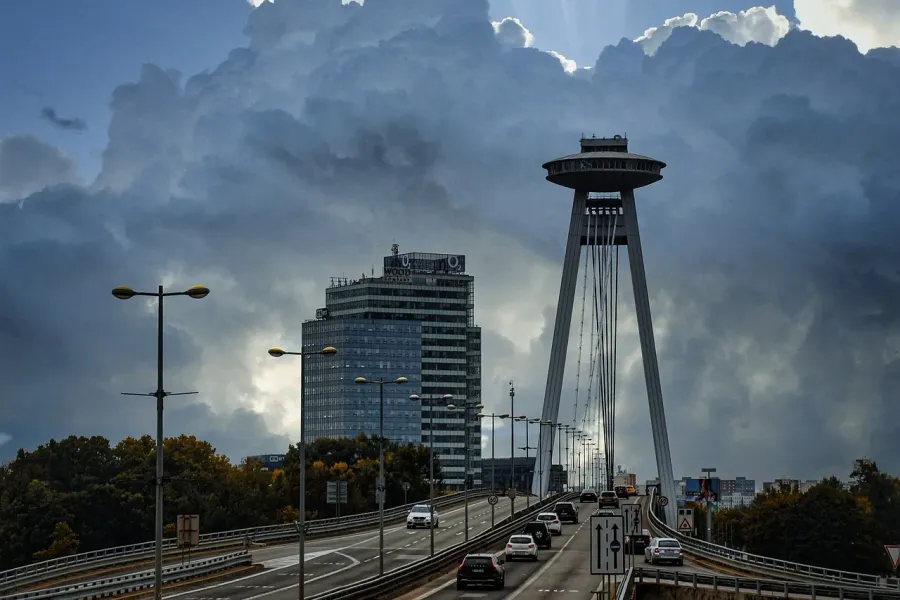 Modern UFO Bridge (Most SNP) in Bratislava with cars on the roadway, a distinctive saucer-shaped restaurant, under dramati...