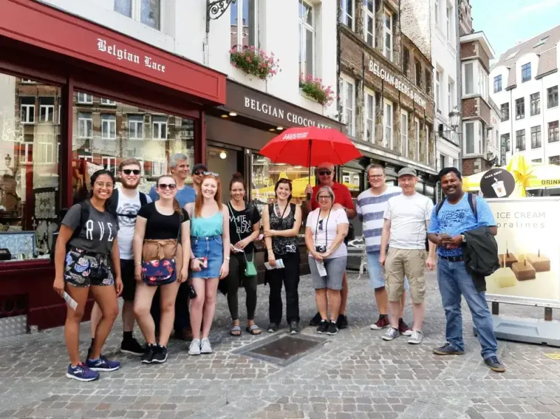 A diverse group of tourists stands smiling on a cobblestone street in front of Belgian Lace and Chocolate shops.
