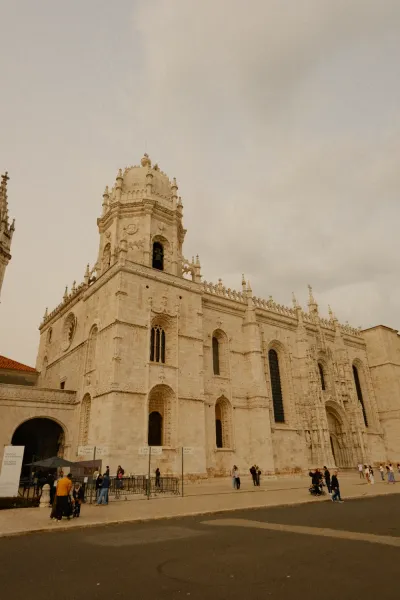 Ornate beige limestone facade of Jerónimos Monastery with a bell tower and arched windows, people walking in front.