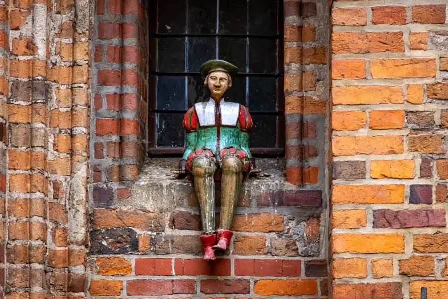 Colorful wooden figure of a man in a green beret and medieval-style clothes, sitting on a brick window sill of an old buil...