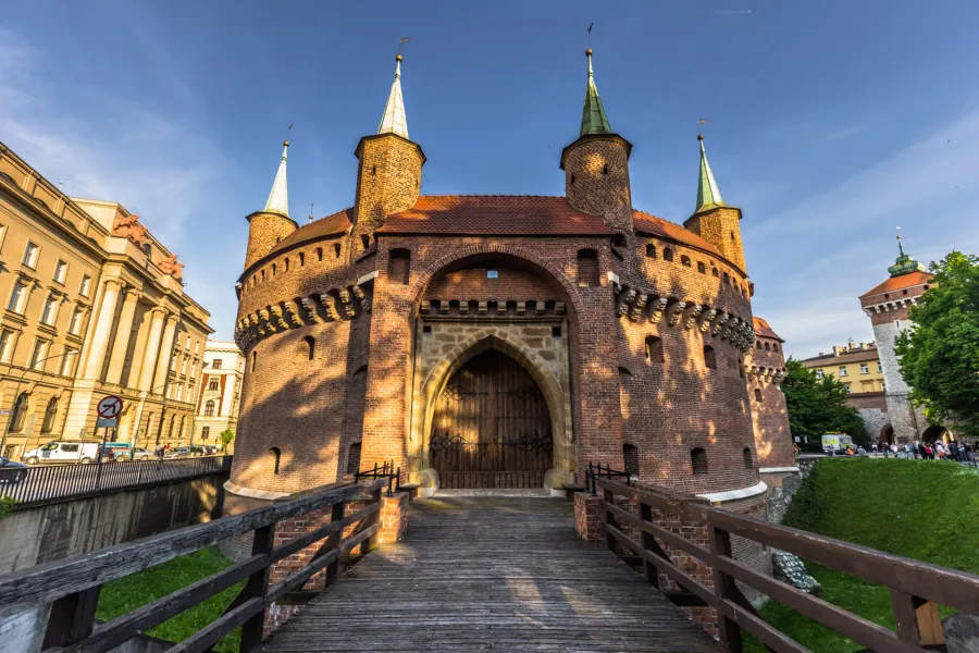 The Krakow Barbican, a circular red-brick fortress, seen from its wooden entrance bridge on a sunny day.