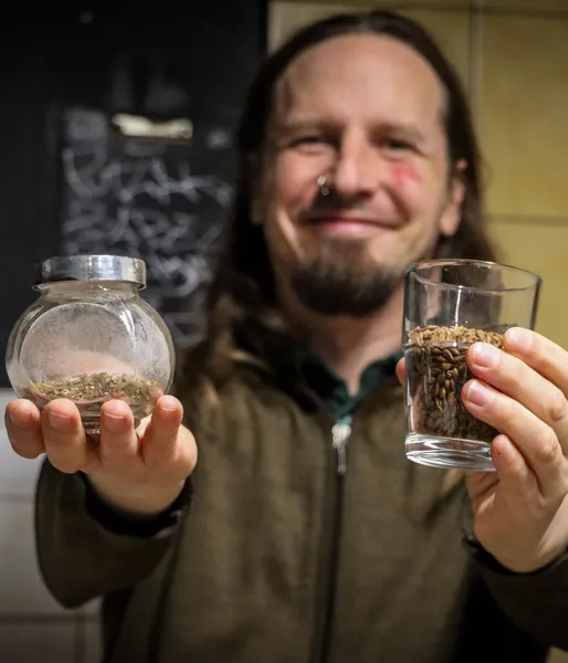 A man with long hair holds up a glass of malted barley and a small jar of dried hops toward the camera.