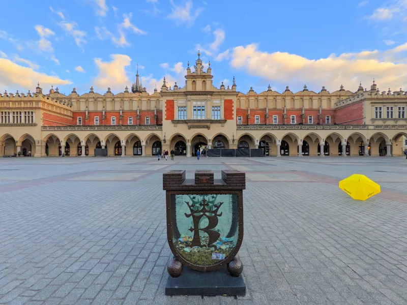 The Cloth Hall in Krakow's Main Market Square at dusk, with a donation box and a yellow umbrella in the foreground.