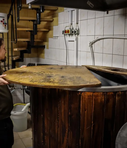 A brewer opens the wooden lid of a large brewing vat in a small brewery.