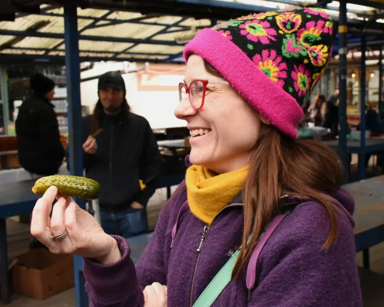 Smiling woman wearing a colorful beanie holding a traditional pickled cucumber at an outdoor food market in Krakow.