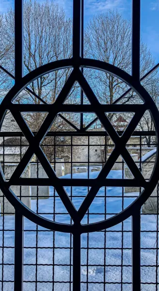 View through a window with a Star of David grille. Outside, a snow-covered cemetery with gravestones and bare trees.