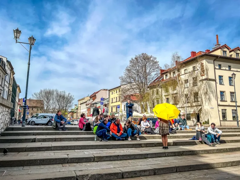 People sit on wide stone steps in a city square, historic buildings and a lamppost under a partly cloudy sky. One person h...