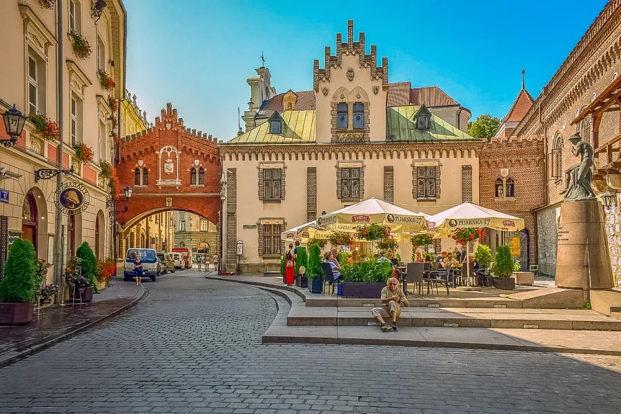 Cobblestone street in Krakow with a red brick archway, historic buildings, and an outdoor cafe under white umbrellas. Peop...