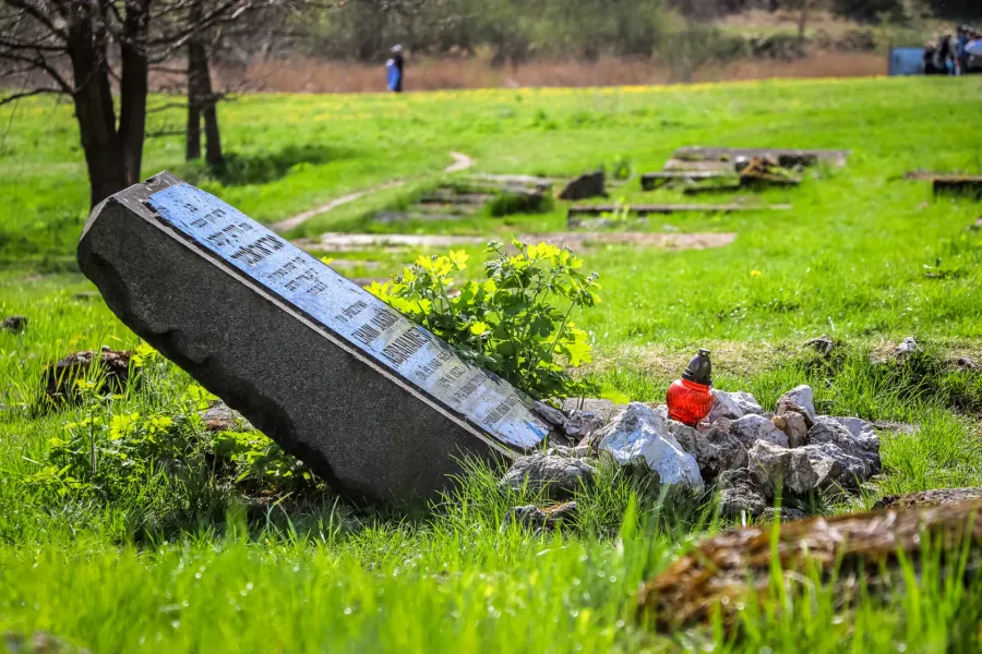 An inclined stone memorial with inscriptions stands in a green field, surrounded by small rocks and a red glass candle.