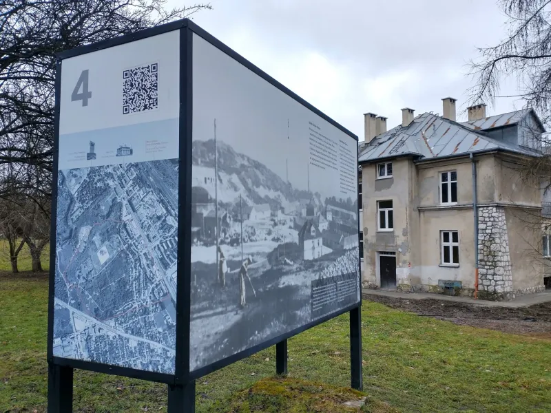 An information board with a map and historical photos stands on a lawn in front of the 'Grey House' at the Plaszow camp site.