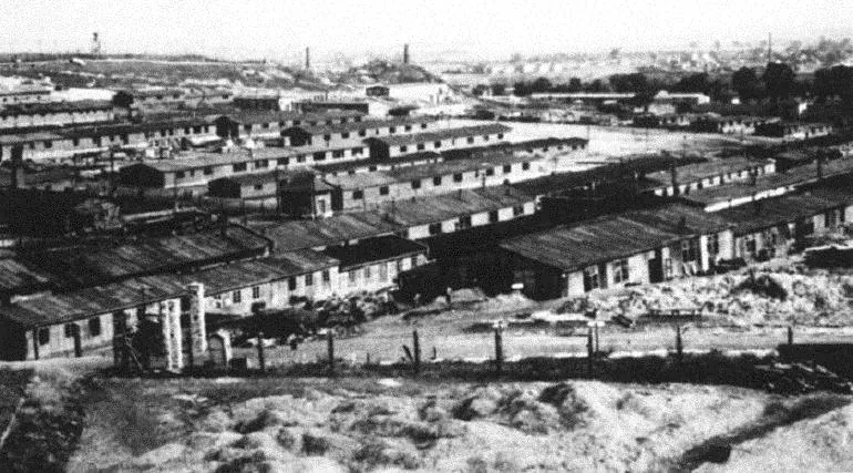 Black and white aerial view of Plaszow Concentration Camp with numerous barracks, fences, and distant industrial structures.