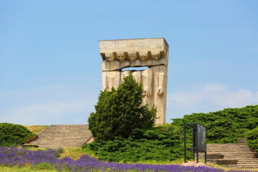 A large, abstract stone memorial monument stands against a clear blue sky, surrounded by green shrubs, stone stairs, and p...