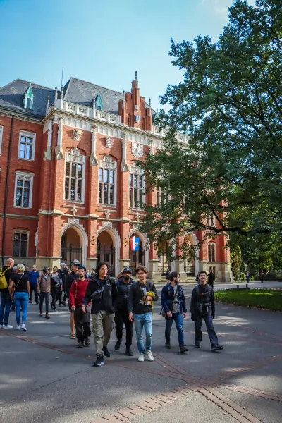 A tour group walks past the red brick neo-Gothic facade of the Collegium Novum in Krakow on a sunny day.