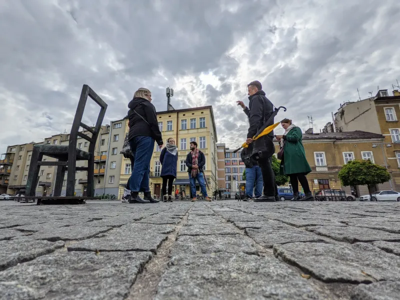 A tour guide speaks to a small group on a cobblestone square with large metal chair sculptures and buildings under a cloud...