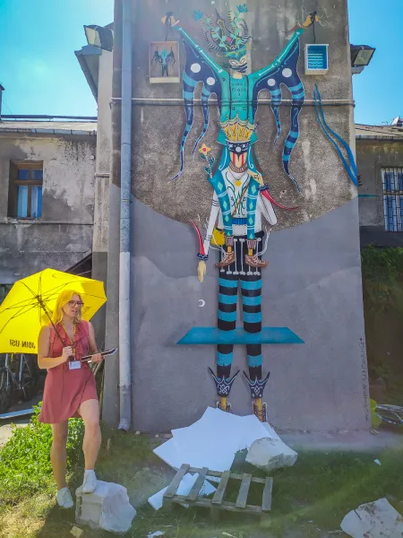 A tour guide with a yellow umbrella stands in front of a large, colorful mural on a building in Krakow.