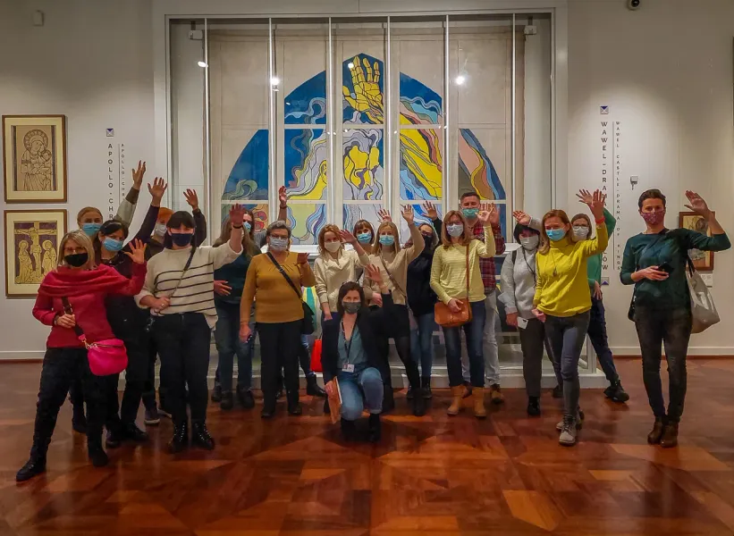 A group of tourists wearing face masks waves inside a museum in front of a large, modern artwork.