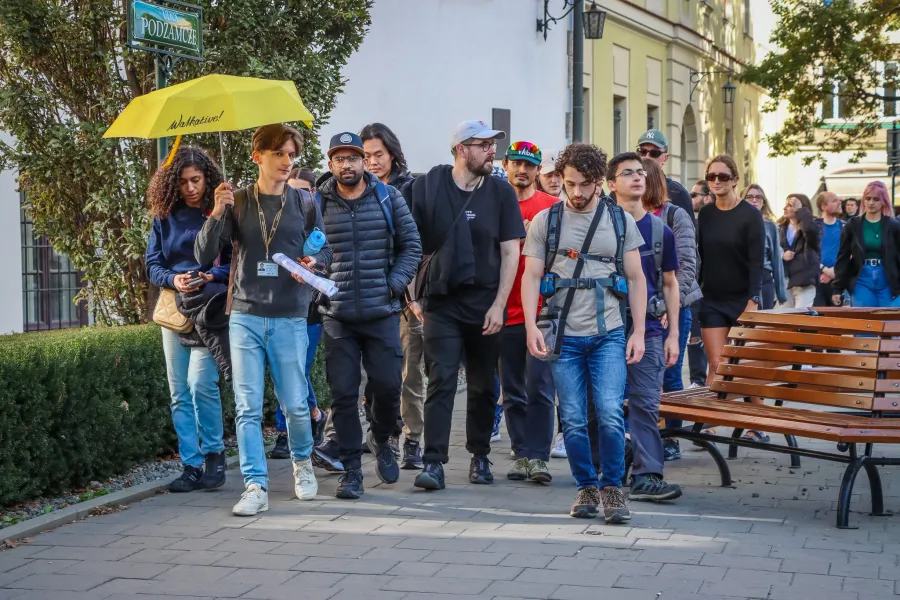 A Walkative tour guide with a yellow umbrella leads a diverse group of tourists along a paved city street in Krakow.