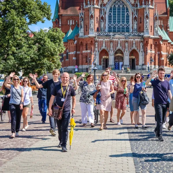 A tour guide with a yellow umbrella leads a group waving at the camera in front of a large brick church in Krakow.