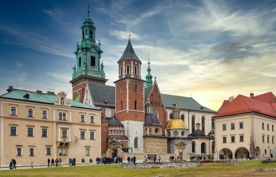 The Wawel Cathedral complex on a partly cloudy day, with its various towers and chapels seen from a grassy lawn.