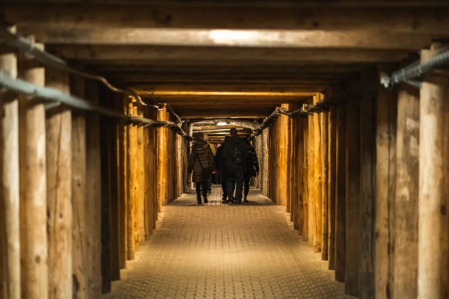 A group of tourists walking through a long, underground corridor supported by heavy wooden beams in the Wieliczka Salt Mine.
