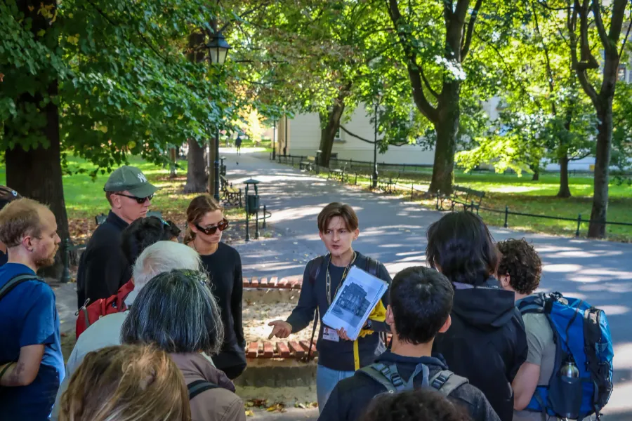 A tour guide shows historical photos to a group of people on a path in a sunlit park in Krakow.
