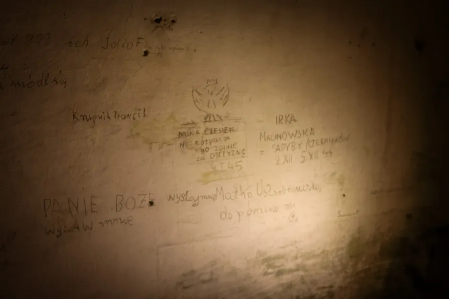 Dimly lit wall with pencil inscriptions in Polish from the WWII era, including a hand-drawn Polish eagle with a crown.