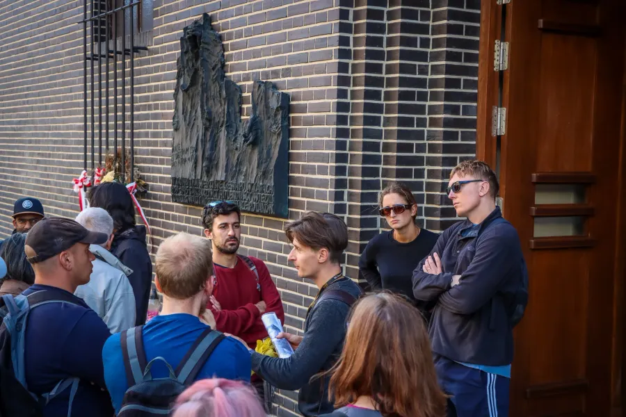 A tour guide explains a point to a group of travellers in front of a memorial plaque on a brick building in Krakow.