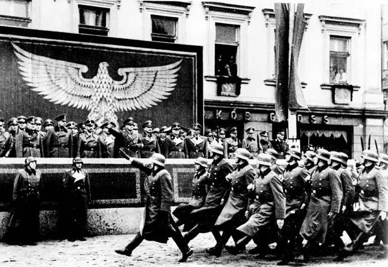 Black and white photo of a Nazi military parade on a street in Krakow. Soldiers march past a stand with officers and a lar...