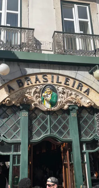 Facade of Café A Brasileira in Lisbon, showing its arched sign, a central relief of a man with a cup, and green-framed win...