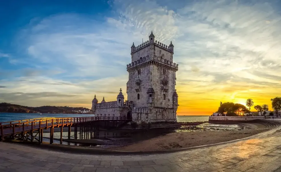 Historic Belém Tower in Lisbon at sunset, with a wooden walkway over the Tagus river and orange-blue sky.