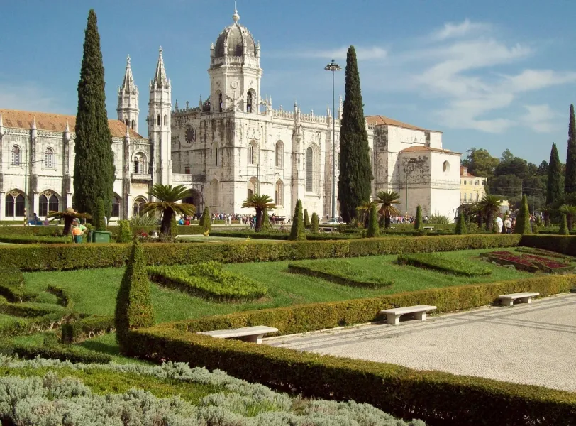 Jerónimos Monastery in Lisbon with manicured green gardens, cypress trees, and visitors on a sunny day.