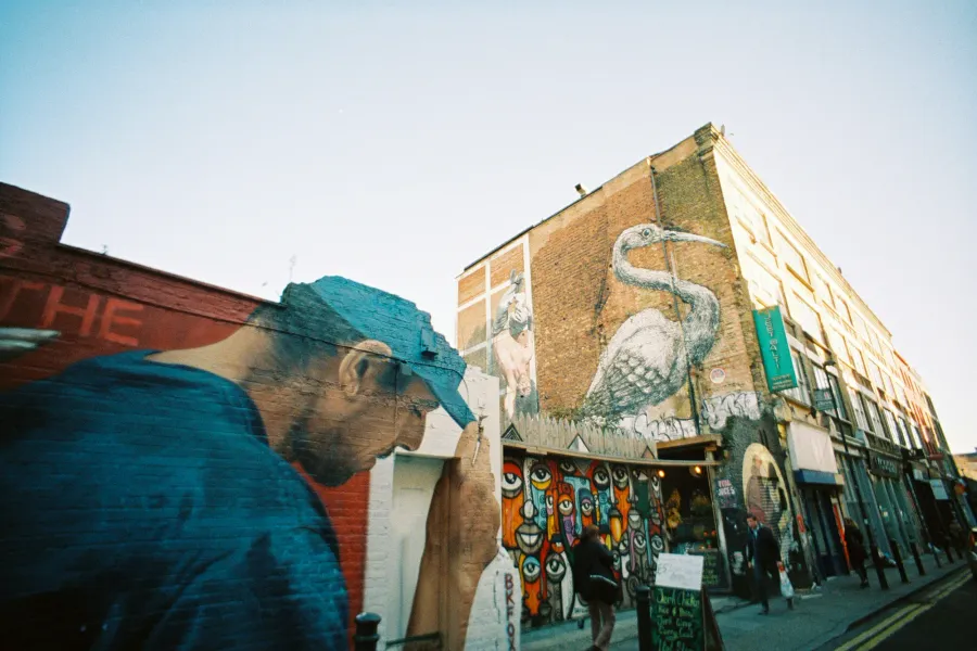 A London street with large-scale murals on brick buildings, including a large white heron and other colourful artwork.