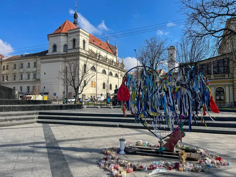 Heart-shaped memorial frame decorated with blue and yellow ribbons and many candles in a Lviv square, with a historic buil...