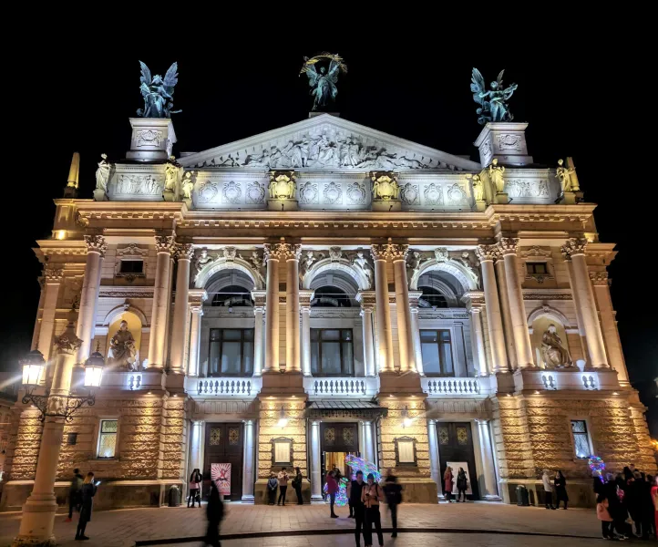 The brightly illuminated Lviv Opera House at night, with detailed sculptures and people walking in front.