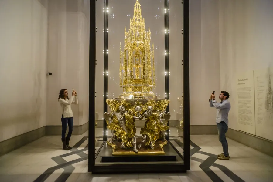 An ornate, gilded monstrance in a glass display case, being photographed by two visitors on their phones.