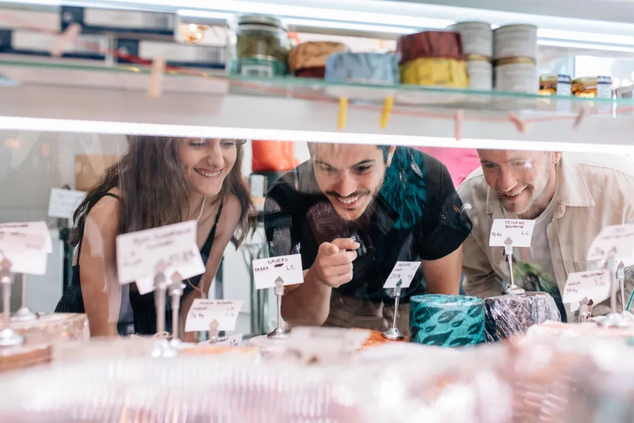 Three young people smile as they look through a glass counter at a selection of cheeses in a shop.