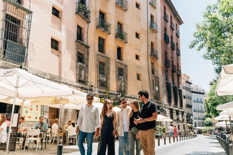 A group of five young people walk down a sunny Madrid street lined with cafe tables and historic buildings.