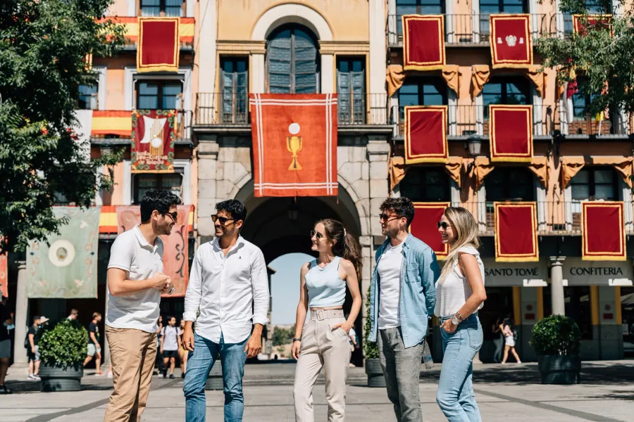 A group of five young adults in a sunny plaza, in front of a building decorated with red and gold banners.