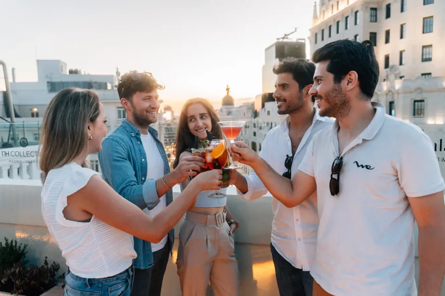 Five young friends toast with colorful cocktails on a rooftop bar in Madrid at sunset.