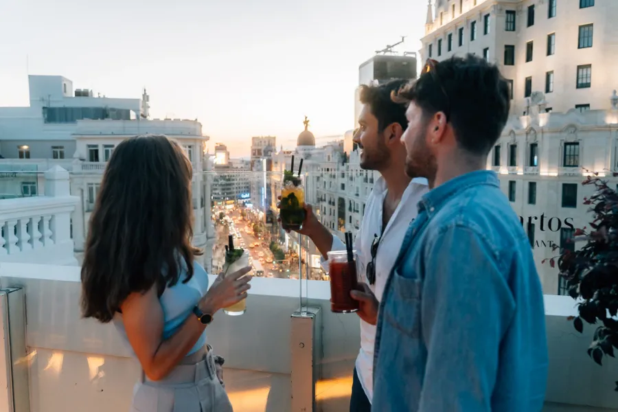 Three friends with cocktails on a rooftop terrace overlooking the busy streets of Madrid at sunset.