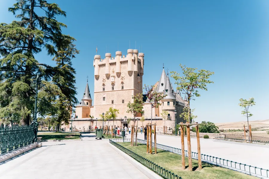 The Alcázar of Segovia, a stone castle with turrets, seen from a park on a sunny day.