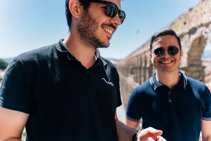 Two young men in sunglasses stand smiling in front of the Aqueduct of Segovia on a sunny day.