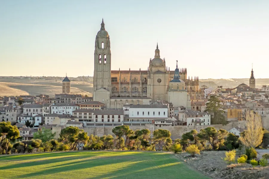The Segovia Cathedral stands over the old town, seen from a green hill at sunset.